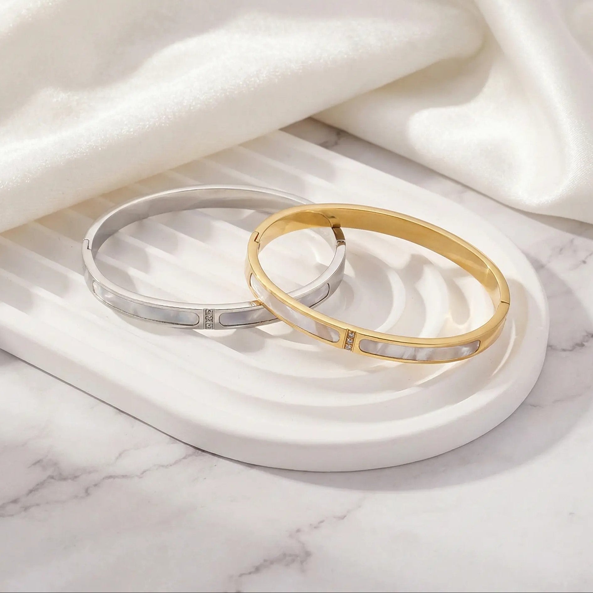 Two bangles, one gold and one silver, on a marble surface with white fabric.